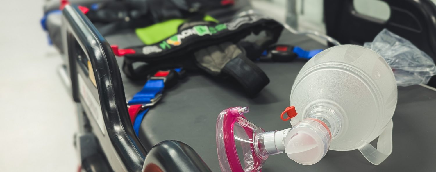 A photo of a bag valve mask with anaesthetic mask attached sitting on top of transport trolley with ACR harness strapped on to trolley mattress