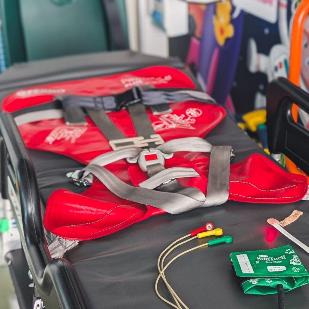 Red Ferno pedimate plus harness secured to paediatric transport trolley with blood pressure cuff and ECG dots on trolley.